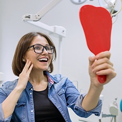 A middle-aged woman wearing glasses and looking at her new smile thanks to affordable dental implants