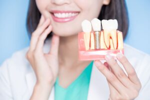Blurry background image of a woman touching her jaw while holding a model of a dental implant in the foreground